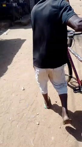 A man is walking down a dusty road, pushing a bicycle with a blue container strapped to the back. He is wearing a black t-shirt, white shorts, and sandals. He is bald. He is walking past a small building with a sign that reads "De Nize Fashion." The sign lists a phone number and the services offered, including suits, caftans, native wear, and hats. The environment is sunlit and appears to be a rural or less developed area. There is some trash on the ground. Another person is walking in the distance.