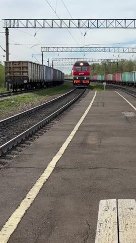 A red and grey electric train approaches the camera on a train track at a train station. The train has the logo “РЖД” on it. The train tracks are black, with yellow painted lines on the concrete. Other trains and rail cars sit on tracks on either side of the track the red train is on. Overhead are cross-wire metal structures and power lines. The video is shot in daylight with an overcast sky.