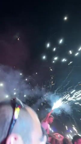 The video shows a concert crowd at night. The sky is black, filled with numerous bright white lights, possibly from lasers or fireworks. A man wearing a white tank top sits on someone's shoulders, holding a lit sparkler high in the air. Sparkles and smoke fill the space around him. The faces of other concertgoers are visible in the foreground, looking towards the man with the sparkler. There are some colorful tents visible in the background.