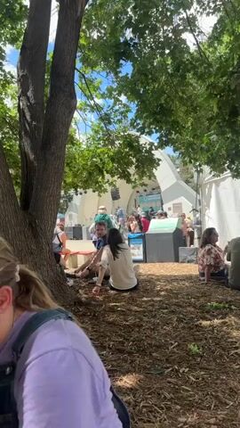 The video shows a live band performing on an outdoor stage, as viewed from behind a crowd of people sitting on the ground. The stage has a white, arched backdrop and a banner advertising "Bands on the Bricks" sponsored by Avery Brewing. The band consists of several musicians playing guitars, a fiddle, and possibly other instruments. The crowd is seated on the ground covered in wood chips, under the shade of a tree. A blue recycle bin is visible in the foreground. People in the crowd are dressed in casual summer clothes. Some are standing near the stage, while others are seated, listening to the music.
