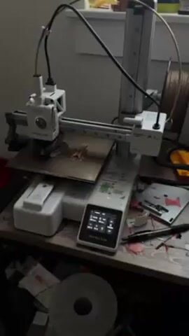 The video shows a Bambu Lab A1 Mini 3D printer printing two small, brown objects on its gold-colored print bed. The printer is mostly white and gray and is located on a cluttered desk next to a window. The desk has various items scattered around, including a spool of filament, packaging, and writing utensils. The printer's touch screen displays settings.  The printer's nozzle moves back and forth over the print bed, depositing filament to build up the objects.