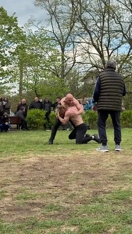 In a park-like setting, two men are wrestling on a patch of grass, surrounded by a crowd of spectators. The wrestler on top is bald and shirtless, wearing black pants and shoes. He is gripping the other wrestler, who is kneeling and also wearing black pants and shoes. An older man in a green vest and dark pants stands nearby, appearing to referee the match. The spectators consist of a mixed group of men and women, some standing and others seated on a bench. The environment includes trees, green grass, and a cloudy sky.