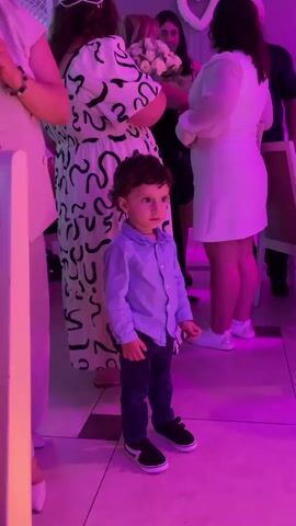 A young boy stands in the foreground of the video, dressed in a light blue button-down shirt and dark pants. He has dark, curly hair and is looking to his right. Behind him, several people are gathered. To the left, a woman in a white suit with a black top is partially visible. To the right of the boy, a woman in a white dress is holding a bouquet of flowers. Further in the background, other individuals are present, and the environment appears to be an indoor event with purple lighting. The floor has a tiled pattern. The video is short, showing slight movement and zooms on the boy.