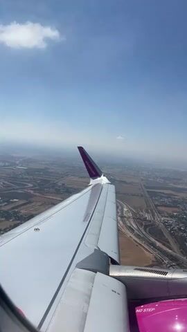 The video shows an aerial view from an airplane window. The right wing of the airplane is prominent in the foreground, with its white surface and purple wingtip. Below the wing, a landscape is visible, featuring agricultural fields, roads, and scattered buildings. The sky is blue with a few wispy clouds. The airplane appears to be in flight, with the ground receding below.