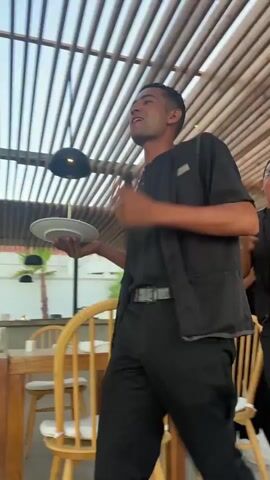 This is a short, horizontal video clip showing a man in a black uniform, likely a waiter or server, as he holds and plays a large frame drum. The man is outdoors, under a wooden slatted roof structure. In the background, there are tables and chairs, suggesting a restaurant or dining area. To the right, glimpses of a beach and the sea are visible, with a sunset in the distance, indicating it is late afternoon or early evening. The man is animated and appears to be enjoying himself, smiling and looking up as he plays the drum. Another person's hand is visible in the background to the right. The overall scene suggests a festive atmosphere, possibly a celebration like a birthday, as the audio includes a snippet of the "Happy Birthday" song. The setting appears to be a resort or hotel in Hurghada, Egypt, given the beach and the warm, sunny climate suggested by the lighting. There is no visible text in the clip.