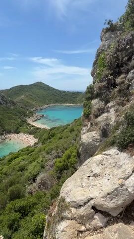 The video pans across a picturesque bay with turquoise waters. Lush green hills surround the bay, which is dotted with small boats and a beach filled with sunbathers. The sky is a clear blue with a few wispy clouds. The camera is positioned high on a rocky cliff overlooking the scene.