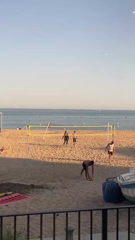 Four people are playing beach volleyball on a sunny day.  There is a yellow volleyball net stretched across the sandy court, with the ocean and horizon visible in the background. Several buoys float in the water, and a few boats are visible in the distance.  A black iron railing is visible at the bottom of the frame, partially obscuring some red and blue objects in the foreground.