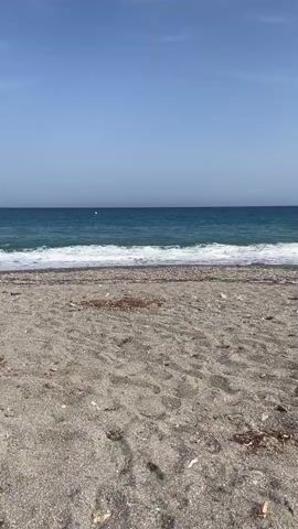This is a static shot of a beach on a clear day. The foreground is a sandy beach with some scattered debris. The midground is the ocean with gentle waves breaking. The background is a clear blue sky with a few faint clouds. There are no people or animals visible in the video.