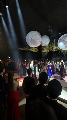 The video shows a dimly lit indoor event space with a large gathering of people, predominantly young adults dressed in formal attire. Three large, illuminated spheres resembling moons are suspended from the ceiling. The ceiling itself is adorned with a canopy of twinkling lights, giving the impression of a starry night sky. Spotlights from above illuminate sections of the crowd and the dance floor area, which has a red and white patterned surface. In the foreground, several individuals are visible from behind, observing the scene. The attendees appear to be mingling and some are walking towards the center of the space where a woman in a light blue, embellished gown is prominent. The overall atmosphere suggests a formal celebration or gala event.