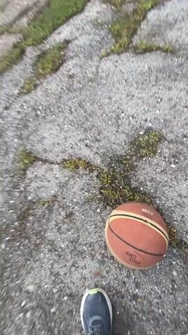 The video shows a first-person perspective of someone playing basketball on an outdoor court. The court appears worn and cracked, with grass growing through the pavement. A basketball hoop with a blue backboard is visible in the background, as are trees and other greenery. The person is dribbling the ball and moving around the court. The footage is shaky and appears to be taken while in motion.