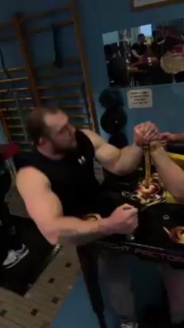 Two men are arm wrestling at a table that is labeled "ARM FIGHT FACTORY" in a room with pale blue walls. One man is wearing a black tank top and gray sweatpants, and has a tattoo on his left arm. The other man is also wearing a black shirt. They are both gripping handles attached to the table with their free hands.  A ladder and some ropes are leaning against the wall in the background.  There is a mirror on the wall reflecting other people watching the arm wrestling match.