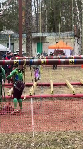 The video shows an outdoor obstacle course.  Two people, one in a green and one in a purple outfit, are at the starting positions, appearing to be hooking themselves up to ropes. There is a crowd behind a red plastic fence, with tents and buildings visible in the background.  Each starting position is marked with a number, and the course involves hanging from ropes. The environment is a somewhat wooded area with leafless trees, suggesting it may be early spring or late fall.