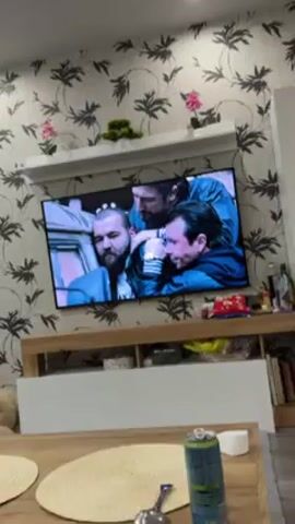 The video shows a television displaying a scene with three men in close proximity, seemingly engaged in a tense or aggressive interaction. The man at the forefront has a beard and a noticeable facial expression of strain or pain. Two other men are either restraining or confronting him. The scene appears to be set in or around a vehicle. The television is mounted on a wall above a wooden shelf unit with decorative objects. The room has a patterned wallpaper with floral and leaf designs. In the lower portion of the frame, there is a glimpse of a table and other miscellaneous items.