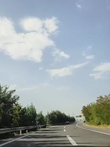 The video shows a view from inside a car driving on a highway. The car is traveling on the right side of the road. There are trees lining the side of the highway, and a guardrail is visible. The sky is blue with some clouds. There are other cars on the highway in the distance. The sun is shining, casting shadows on the road. There is a highway sign visible in the distance.