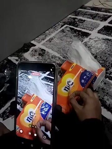 The video shows a person holding a phone to record a stack of tissue boxes. The tissue boxes are orange and blue with a yellow logo that looks like a heart. A few tissues are pulled out of the top box. The person’s hands are visible. The phone screen shows a smaller image of the scene being recorded. The background is a black and white patterned surface. There is a power cable to the right in the background.