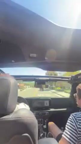 The video shows a car ride on a sunny day, with the camera positioned inside the car looking out through the partially open sunroof. Two people are visible: the driver and a passenger wearing a striped shirt. The car drives on a street lined with houses and trees. At one point, the car passes a gas station. The sky is clear and bright.