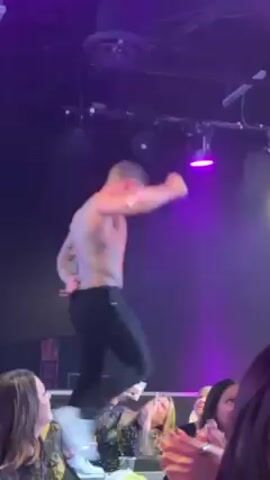 A shirtless man with tattoos dances onstage at a nightclub in front of a crowd of female fans. The man is wearing black pants and moving his arms, showing off his biceps. The stage is bathed in purple light, and there are sound and lighting equipment overhead. A few women in the crowd appear very excited.