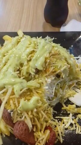 A close-up shot shows a plate of food on a wooden surface. The food appears to be a mixture of thin-cut french fries, possibly chorizo or sausage slices, and a light green sauce drizzled generously over the top. Some white crumbles, possibly cheese, are also visible. The lighting is somewhat diffused. The plate is black. A dark object is partially visible in the upper left corner of the frame.