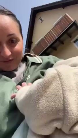 A woman is sitting outdoors, holding a baby. The woman is looking to the right of the frame with a slight smile. She has dark hair pulled back and is wearing a green collared shirt over a grey shirt. The baby is wrapped in a cream-colored fleece blanket. Behind the woman is a two-story house with a tan exterior, a dark roof, a small window, and a wooden balcony. The sky is blue and clear.