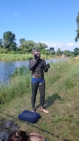 A boy is covered in mud, standing on the grassy bank of a river. He is wearing blue shorts. He is smearing mud on his face and chest. To his left is an inflatable pillow. In the foreground, a person wearing goggles is lying face down in the mud. The river is calm and reflects the blue sky. Trees and reeds line the opposite bank. The overall environment appears to be a natural, outdoor setting on a sunny day.