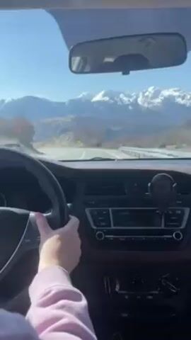 The video shows a person driving a car on a winding mountain road. The person is wearing a light pink shirt. The car has a black steering wheel, a brown dashboard, and a phone mounted on the dashboard. The rearview mirror shows the road behind the car.  Outside the car are mountains with snow-capped peaks. The sky is clear and blue.  A road sign for a sharp left turn is visible on the side of the road.  The interior of the car is clean.