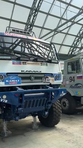 This media file features a close-up, low-angle shot of a large, white Kamaz Dakar rally truck. The truck is predominantly white with blue accents and prominent sponsorship decals. The Red Bull logo is visible at the top of the windshield, and other stickers include "Macro Total," "ARGENTINA BOLIVIA CHILE," "SpeedAgro," "Team Kamaz master," and the number "507" within a Dakar logo. The truck's massive, knobby tires suggest it is designed for off-road racing. The front bumper is a heavy-duty blue metal structure. The vehicle is indoors, under a modern, industrial-style ceiling with visible metal beams and translucent panels that allow light to filter through, suggesting daytime. Two of the front tires are elevated on supports, indicating maintenance or display. In the background, another truck, partially visible, sports the number "94" and a different set of logos. The overall scene appears to be a garage or exhibition space, possibly at a rally team's facility or a museum, given the preserved state of the truck. No people are clearly visible, though a hint of a person can be seen in the far background to the left. The setting is likely Verkhnyaya Pyshma, Russia, as indicated by the provided location context. The lighting is bright, consistent with daylight.
