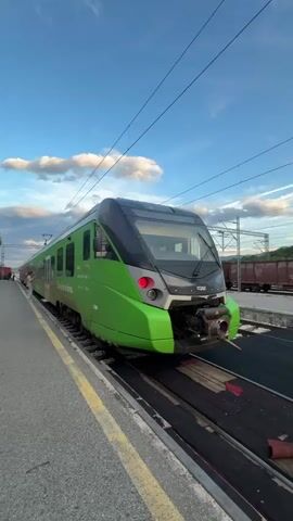 The video shows a green train with the text "CAF" on its side, sitting on train tracks at a station. The train station building, made of light-colored stone, is behind the train and to the left. The sign on the building reads "НИКШИЋ-NIKŠIĆ". A flag hangs from the building. There are benches along the platform, where people are sitting. The sky above is blue with scattered clouds.