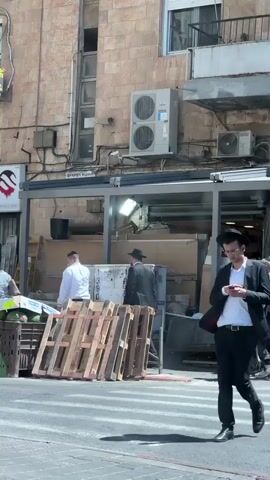 The video shows a street scene with several men, some of whom are dressed in traditional Orthodox Jewish attire. In the foreground, two men are walking on a pedestrian crossing. The man on the left is wearing a black suit, a white shirt, a black hat, and a tallit. The man on the right is wearing a black suit, a white shirt, and a black hat and is looking at his phone. In the background, there are buildings with a brick facade. There are also air conditioning units mounted on the buildings, electrical wires, and scaffolding. Several wooden pallets are stacked in the street, and there appear to be some crates or boxes behind them. One man in the background is wearing a white shirt and speaking to another person. Another man is wearing a dark suit and hat and is standing near the pallets. The scene suggests a busy urban environment, possibly in a city with a significant Orthodox Jewish population.