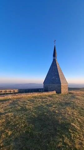 The video shows a small stone chapel with a dark, pointed roof and a cross on top. The chapel is situated on a grassy hilltop under a clear blue sky. The sun is low on the horizon, casting a warm glow and creating lens flares across the scene. In the distance, a hazy landscape can be seen, with some structures or landmarks visible on the horizon. The grass on the hill is dry and golden in places, suggesting it might be late autumn or winter. The camera pans slowly to the right, revealing more of the sunlit landscape and a faint silhouette of another structure on the right side of the frame. There are no people or animals visible in the video.