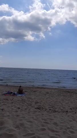 The video shows a cloudy sky above a body of water and a beach. In the foreground on the sand, a person in a dark swimsuit sits on a towel. The person is facing away from the camera, looking out at the water. Further out in the water, two people can be seen swimming. The sun is partially visible through a break in the clouds, casting a bright spot in the sky.