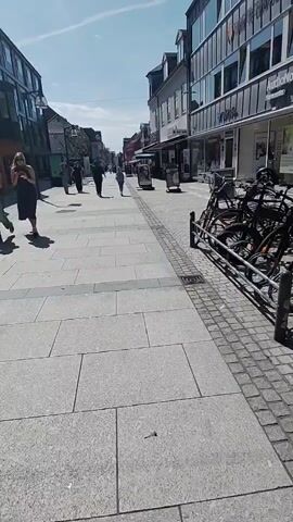 This is a first-person perspective video of a city street. The camera is moving forward, showing a paved sidewalk with large gray stone slabs and a textured gray border with a grate. On the right side of the sidewalk, there is a metal bike rack with several bicycles parked. Behind the bike rack are storefronts with windows and awnings, one of which has the name "AudioNova" visible. People are walking in the distance on the sidewalk, moving away from the camera. The street is lined with buildings, and the sky is blue with some clouds. The lighting suggests it is daytime.