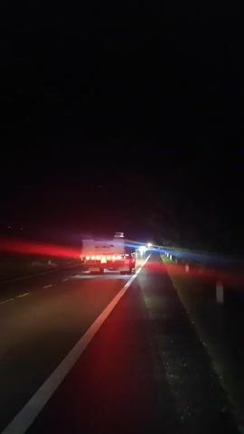 The video shows a view from inside a moving vehicle at night. The road ahead is visible, with lane markings illuminated by headlights. Several vehicles are ahead, including trucks, whose taillights and hazard lights cast red and white reflections on the wet road surface. The sky is dark, indicating it is nighttime. There are trees or foliage visible on the sides of the road. The camera appears to be mounted at a high vantage point, looking forward and slightly down. The video is shaky, suggesting the vehicle is in motion.