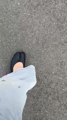 A person wearing black split-toe shoes and light gray pants with paint splatters walks on asphalt. The camera is positioned directly above the person’s feet, focusing primarily on the shoes and pants. The asphalt surface is visible as the background. The person moves forward at a steady pace.