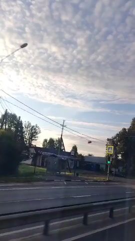 The video shows a view from a moving vehicle looking out the side window. The scene is a road with trees and buildings on the side. There are streetlights and pedestrian crossing signs visible. The sky is partly cloudy with the sun shining through, creating lens flares and glare on the window. The road appears to be a highway or a main road with a metal guardrail in the foreground. The overall impression is a sunny day in a suburban or rural area.