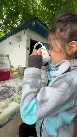 This is a vertical, close-up video clip of a young girl sitting at an outdoor table. She is wearing a grey and blue patterned jacket and has dark hair. In the foreground, she holds a white mug with a black bird pattern and drinks from it. To her left, a large glass jar filled with a red liquid sits on the table, along with another grey mug. The table is covered with a cloth featuring a floral print. In the background, a small white building with a blue-trimmed roof is visible, and greenery suggests an outdoor, possibly rural, setting. The lighting suggests it is daytime, and the overall atmosphere appears relaxed and casual. The girl looks towards the camera with a slight smile at the end of the clip, conveying a sense of contentment or curiosity.