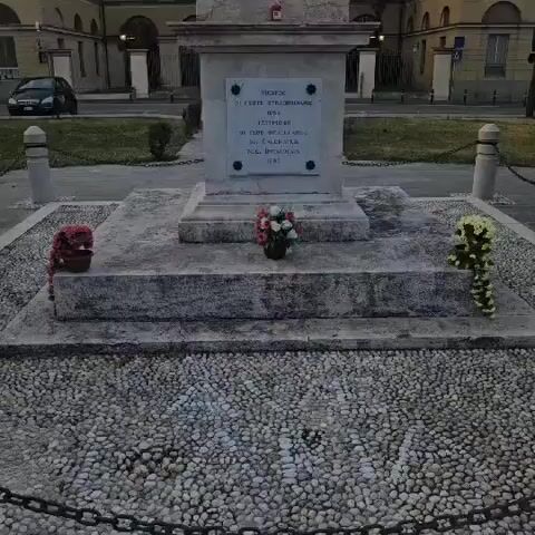 A video captures a monument in an outdoor setting. The monument features a large white column topped by a pedestal and a statue of a woman with a halo. The base of the monument is adorned with wreaths and flowers. In the background, a building with a classical architectural style is visible, along with streetlights and a car parked on the side. The sky appears partly cloudy. The video pans up the monument, focusing on its details and the statue at the apex.