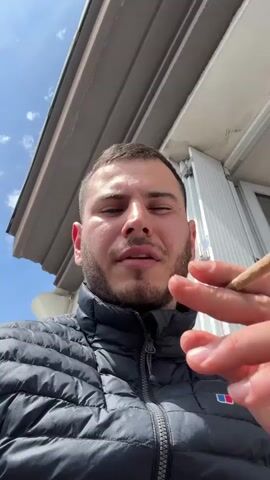 A light-skinned man is seen in a selfie-style video, holding and smoking what appears to be a hand-rolled cigarette. Initially, the man is wearing a black, quilted down jacket. He then removes the jacket, revealing a black, quilted down vest. The man has short, dark hair, a light beard, and is looking directly at the camera. The background shows a partially cloudy sky and the eaves of a building, which transitions to a view of an urban streetscape from what appears to be a balcony. The street scene includes cars, buildings, and a glimpse of greenery. The general impression is of a sunny day in a European city.