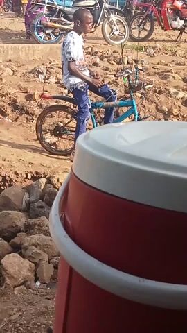 A man, dressed in a white patterned shirt and blue jeans, is lying on his back on a blue bicycle. Several other bicycles and motorcycles are parked nearby. The ground appears to be uneven dirt with some rocks. A large, cylindrical red and white object occupies part of the right side of the frame, obscuring some of the background. The overall scene appears to be outdoors, possibly in a market or parking area.