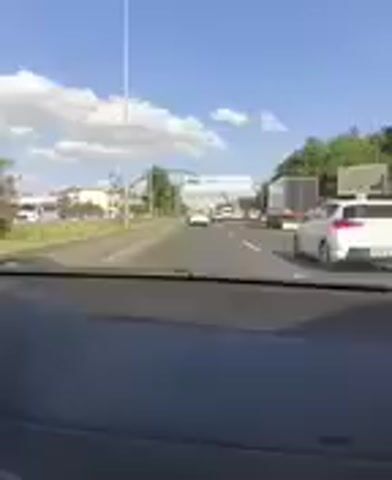 This media file captures a view from a vehicle driving on a highway, likely in Mamak, Turkey, given the context. The scene depicts moderate traffic on a sunny day with scattered clouds in a bright blue sky. The perspective is from the driver's seat, looking forward through the windshield.

The road is multi-lane, with several cars, vans, and at least one truck visible. The vehicles are moving, but the presence of a traffic jam is implied as they appear to be in slow-moving lanes. On the left side of the highway, there is a grassy median strip and some trees. In the distance, there are buildings and what appear to be further trees, suggesting an urban or suburban environment.

A prominent feature of the scene is a large, modern pedestrian bridge spanning the highway. The bridge has a distinctive arched structure supported by vertical pillars. Above the roadway, there are also overhead signs. One sign clearly displays directional information, with blue and white text. Though pixelated, some letters are decipherable on the blue section, possibly indicating "Şehitlik" and "Kazım Karabekir," common place names in Turkey. Another sign to the right appears to be a white information board with some text and symbols, possibly arrows indicating lane usage.

The time of day appears to be daytime, likely late morning or early afternoon, given the bright sunlight and the shadows cast. The weather seems pleasant and clear, ideal for travel. There are no people clearly visible within the vehicle or on the side of the road, and the overall mood conveyed is that of everyday commute or travel. The focus is on the road ahead and the surrounding infrastructure, capturing a typical highway scene in Turkey.