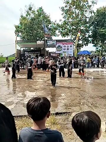 The video shows a group of people performing in an outdoor, public setting, likely a cultural event or festival. The performance is taking place on a wet, possibly flooded, paved area. A band appears to be playing on a makeshift stage behind the performers. There's a crowd watching, and some people are moving around the performance area. Performers are wearing similar dark clothing, and some are carrying sticks or rods as part of their routine. There are trees and some flags in the background. The weather appears to be overcast or partly cloudy.
