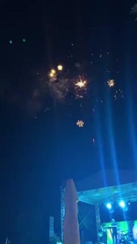 The video shows a nighttime scene with fireworks exploding in the sky. Below the fireworks, there is a stage with bright blue lights and several figures that appear to be musicians. A large, pale yellow object, possibly an umbrella or tent, is positioned in the foreground, obscuring part of the stage. The sky is dark, indicating it is night, and the fireworks are the main source of light and color in the upper portion of the frame. The overall atmosphere appears festive.