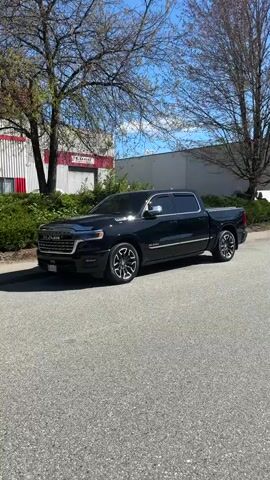 The video shows a black Ram pickup truck parked on a paved road. The truck has chrome accents and custom wheels. Behind the truck, there's a building with a red and white facade and green landscaping. There are also trees with bare branches on either side of the truck. The sky is blue with scattered clouds. The scene is well-lit and appears to be outdoors during the daytime.