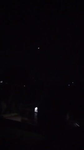 The video shows a dark night sky with a crescent moon visible. Below the sky, there's a dark and indistinct foreground showing hints of trees and possibly buildings, suggested by faint lights. A brighter light reflects off what appears to be water in the lower part of the scene. The camera is static throughout the video.