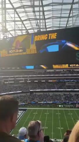 The video shows an American football stadium, likely SoFi Stadium, during a game. The stadium is crowded with fans, and a large screen displays the text "BRING THE THUNDER" along with decibel level readings that fluctuate. There are glimpses of people in the foreground. The video appears to be taken from a spectator's perspective.