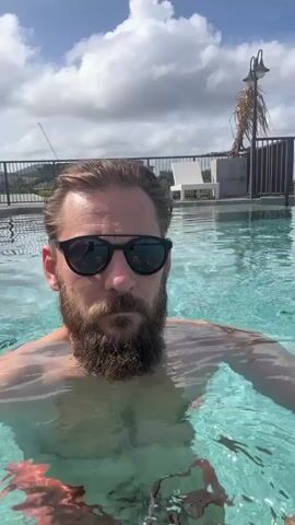 A man with blonde hair, a beard, and sunglasses swims in an infinity pool on a sunny day. The man is facing the camera and appears to be recording a selfie video. In the background, there is a rooftop with lounge chairs and a palm tree. The sky is cloudy.