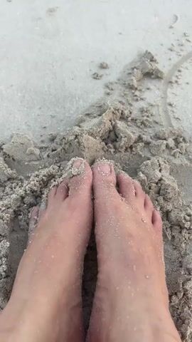 The video begins with a close-up shot of a person's bare feet partially buried in wet sand on a beach. The sand is damp and speckled with small shells and grains. The camera then slowly pans upwards, revealing a wider view of the beach. Gentle waves lap at the shore in the background, and the sky is overcast, creating a soft, diffused light. Footprints are visible in the wet sand leading away from the water. The overall impression is one of a peaceful and serene beach scene.
