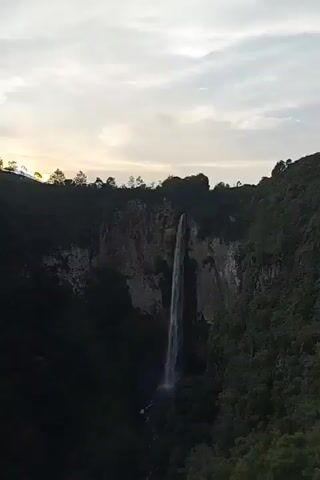 A drone shot pans down from a sky with clouds to reveal a tall waterfall cascading down a rocky cliff face. The waterfall is surrounded by lush green trees and vegetation. The sun is setting, casting a warm glow on the scene. The waterfall is the central focus of the video, with its water flowing in a thin, continuous stream down the cliff. The drone moves closer to the waterfall, providing a more detailed view of the water as it plunges into the pool below. The video ends with a shot of the base of the waterfall, where the water has created a misty spray.