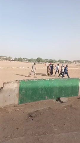 The video depicts a group of approximately seven men walking across a sandy field. They are walking from left to right across the frame. The men are wearing a variety of clothing including traditional robes, white t-shirts, and jackets. The field is bordered by a low concrete wall with a green stripe along the top. In the background, there are rows of tires lined up, presumably marking the boundaries of a track or field. The sky is a pale blue with some haze, suggesting a sunny day. The overall environment appears to be an outdoor recreational area.