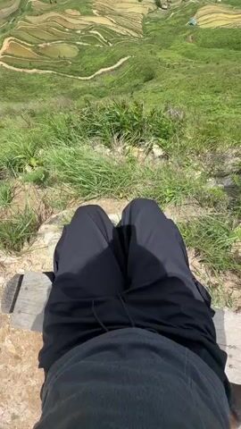 The video begins with a point-of-view shot looking down at a person's legs and lap, wearing black pants and a grey shirt, sitting on a wooden bench. Behind them, a lush green landscape with terraced fields and mountains stretches out under a cloudy sky. A black dog with tan markings sits to the right of the person. The camera then pans to show a wider view of the mountainous terrain, featuring vibrant green hills and winding paths. Several people are seen walking along a dirt path on the edge of a steep incline, with more terraced hills in the background. The sky is filled with dramatic clouds.
