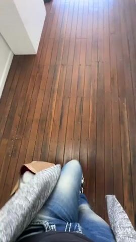 The video shows a first-person perspective of someone sitting in a chair with their legs crossed and wearing blue jeans and white and black sneakers. They are wearing a gray cardigan over a black t-shirt. The floor is made of dark wood planks. To the left, a white surface is visible, and behind it, a black object, possibly a floor lamp, is seen. As the video progresses, the person’s legs shift, and a brown paper bag filled with orange and red items is placed on the floor near their feet. The perspective then shifts to show the person standing and walking towards an open doorway. The doorway leads to an outdoor area with a black mat on the floor. The person is seen from behind, walking through the doorway.